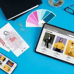 Na ekranie tabletu i telefonu wyświetla się sklep internetowy z ubraniami. Widać też szkice modelek, próbnik kolorów i przybory krawieckie. To inspiracja do stworzenia nowej kolekcji, która może pojawić się jako **okno popup** w sklepie.