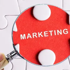 Pod lupą widać słowo "MARKETING" na czerwonym tle, otoczone białymi elementami układanki. To klucz do zrozumienia definicji marketingu.