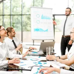 Zespół omawia strategię marketing internetowy. Prezentacja danych na tablicy, ludzie przy stole z dokumentami i laptopem.