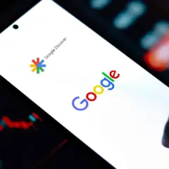 Smartfon z logo Google i napisem "Google Discover". W tle widać wykresy finansowe, sugerujące przeglądanie treści w Google Feed.