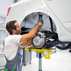 Mechanik naprawia amortyzator w białym samochodzie. To jego praca, a **aso skrót** oznacza, że robi to szybko i sprawnie.