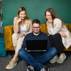 Trzy osoby pracują przy laptopie. Dwie kobiety siedzą na kanapie, a mężczyzna na podłodze. Zastanawiają się, cro co to jest.