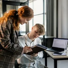 Dwóch młodych ludzi analizuje dokumenty, planując strategię multi channel marketing. Na biurku laptop, telefon i notatnik.