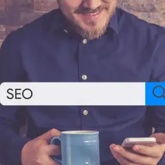 Mężczyzna z kubkiem kawy i telefonem szuka w internecie informacji o SEO. W pasku wyszukiwania widnieje hasło "SEO", sugerujące poszukiwanie w katalogu stron.