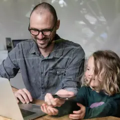 Ojciec z córką przy laptopie, pokazujący jej coś na telefonie. Usługa zdalna.