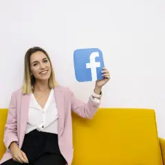 Uśmiechnięta kobieta na żółtej kanapie trzyma niebieski symbol Facebooka. To idealny przykład na marketing szeptany w social mediach.