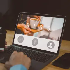 Osoba z kubkiem kawy pracuje przy laptopie, na ekranie którego widnieje napis "BLOG" i ikony.