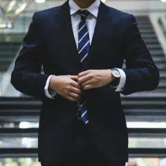 Elegancki mężczyzna w garniturze, zapięty pod szyją, z zegarkiem na ręku, gotowy do roli ambasadora marki.