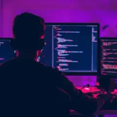 Programista w słuchawkach pracuje przy trzech monitorach, na których widać kod. Wśród linii kodu można dostrzec operatory arytmetyczne.