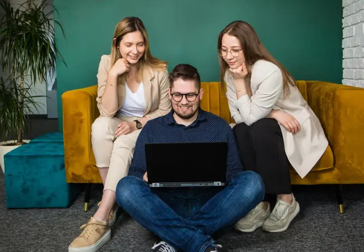 Trzy osoby pracują przy laptopie. Dwie kobiety siedzą na kanapie, a mężczyzna na podłodze. Zastanawiają się, cro co to jest.