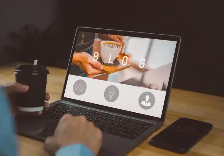 Osoba z kubkiem kawy pracuje przy laptopie, na ekranie którego widnieje napis "BLOG" i ikony.