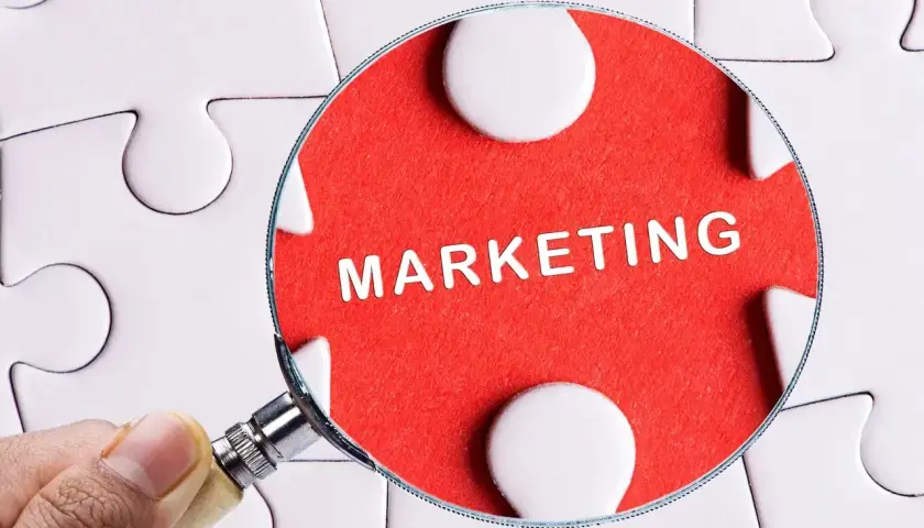 Pod lupą widać słowo "MARKETING" na czerwonym tle, otoczone białymi elementami układanki. To klucz do zrozumienia definicji marketingu.