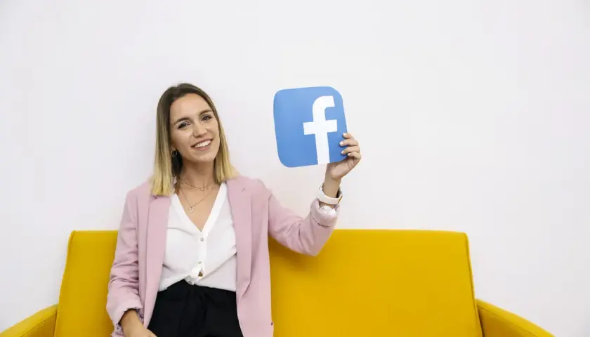 Uśmiechnięta kobieta na żółtej kanapie trzyma niebieski symbol Facebooka. To idealny przykład na marketing szeptany w social mediach.
