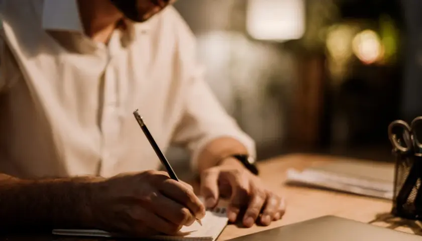 Mężczyzna w białej koszuli pisze ołówkiem w notatniku. Tworzy wartościowy branded content, pracując nad ważnym projektem.