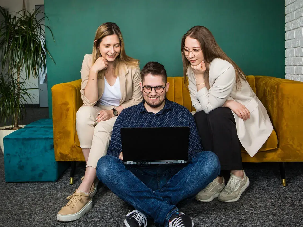 Trzy osoby pracują przy laptopie. Dwie kobiety siedzą na kanapie, a mężczyzna na podłodze. Zastanawiają się, cro co to jest.