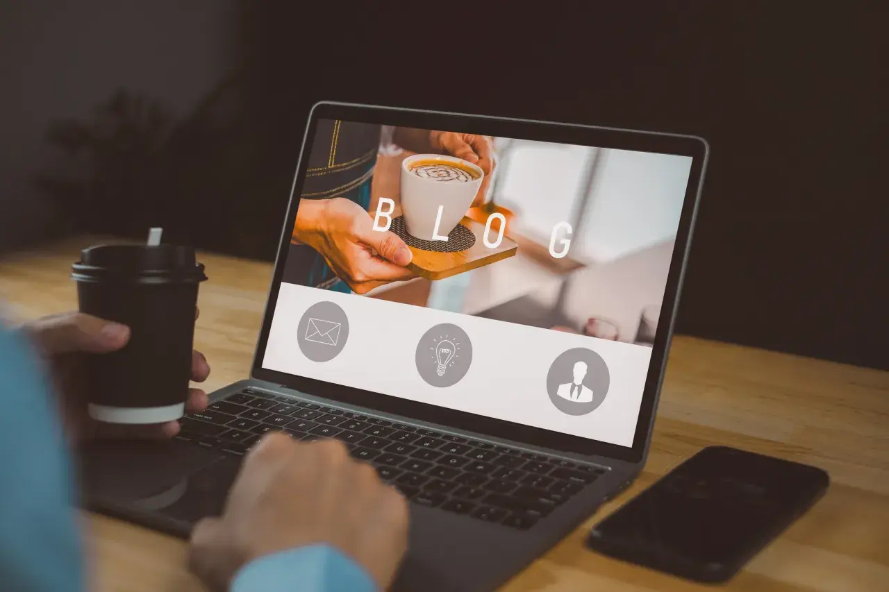 Osoba z kubkiem kawy pracuje przy laptopie, na ekranie którego widnieje napis "BLOG" i ikony.