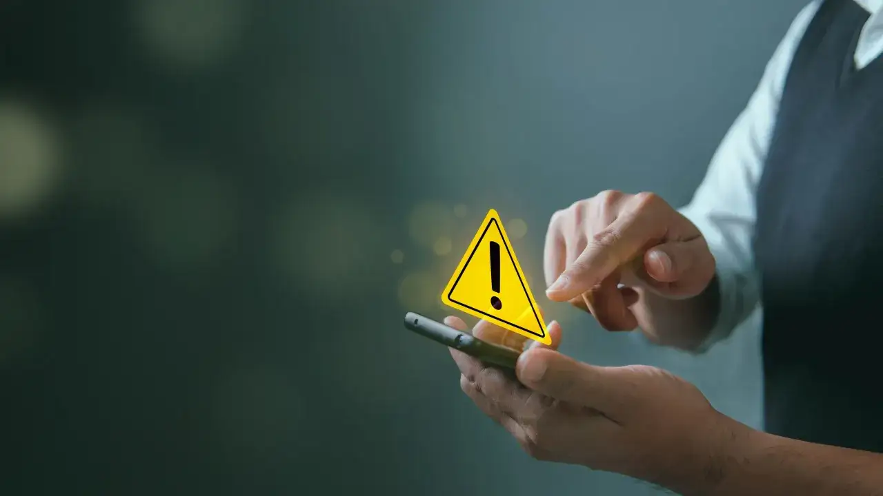 Dłoń trzyma smartfon, na ekranie którego wyświetla się żółty trójkąt z wykrzyknikiem. To ostrzeżenie, co znaczy spam, może być niebezpieczne.