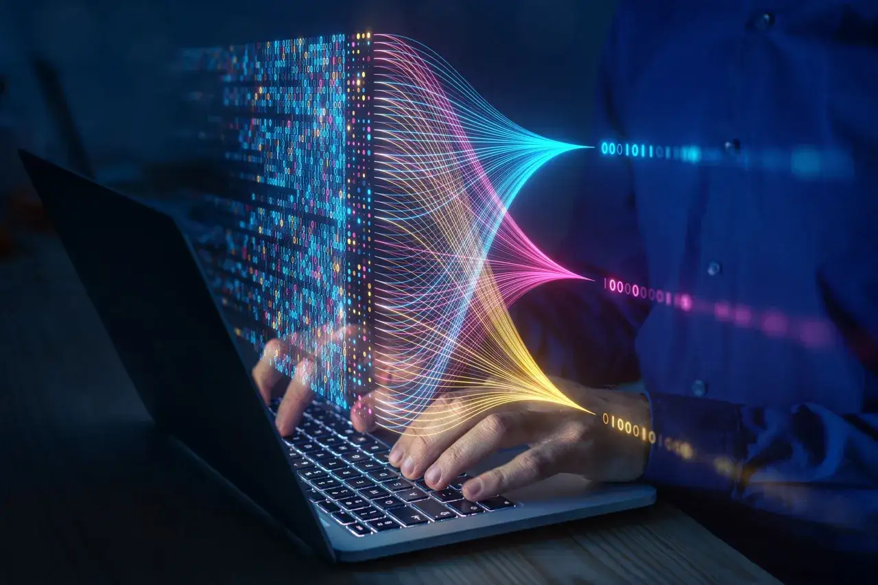 Ręce na klawiaturze laptopa, z ekranu wypływają kolorowe linie danych, wizualizując działanie algorytmu.